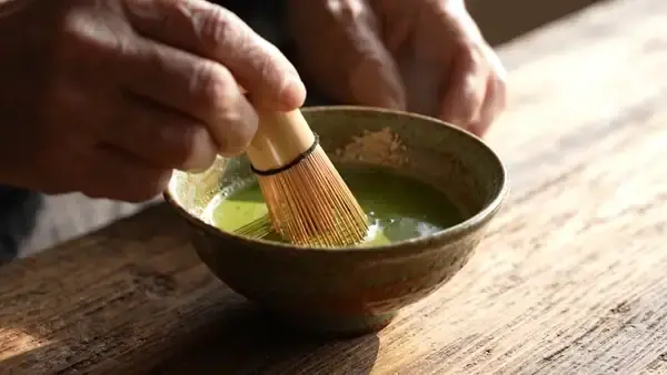 A bamboo whisk moving in a shallow bowl of bright green matcha on a wooden table