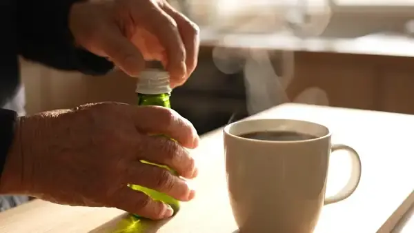 A man's hands twisting the white cap off a small green bottle at a kitchen counter beside a mug of coffee
