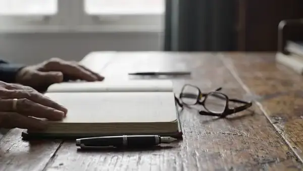 A man's hands closing a ruled accounts book on a desk with a fountain pen laid across the cover